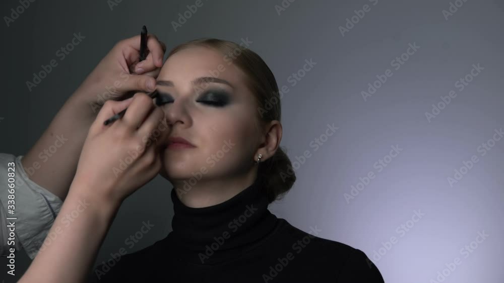 Side view of makeup artist making professional make-up for young woman in beauty studio. Make up Artist makes the makeup smokey eyes of a black shade with special brush of a beautiful model Side view of makeup artist making professional make-up for young woman in beauty studio. Make up Artist makes the makeup smokey eyes of a black shade with special brush of a beautiful model
