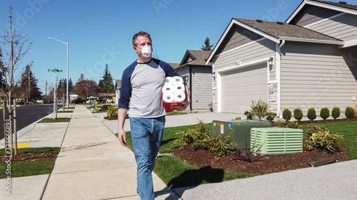 Mature adult male walking alone on an American suburban sidewalk, carrying a pack of toilet paper while wearing a face mask; COVID-19 (novel caronavirus) theme  photo
