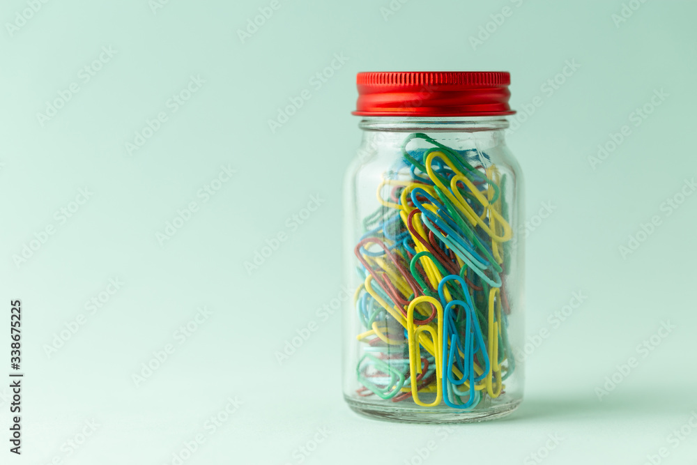 A small glass jar with a red lid full of colorful paperclips on a green background A small glass jar with a red lid full of colorful paperclips on a green background