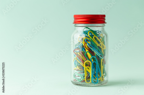 A small glass jar with a red lid full of colorful paperclips on a green background