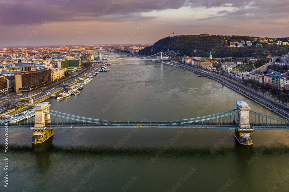 Budapest, Hungary - Aerial view of totally empty Szechenyi Chain Bridge at sunset with Elisabeth Bridge, Citadel and colorful sky at background. No traffic on the bridge due to Coronavirus outbreak