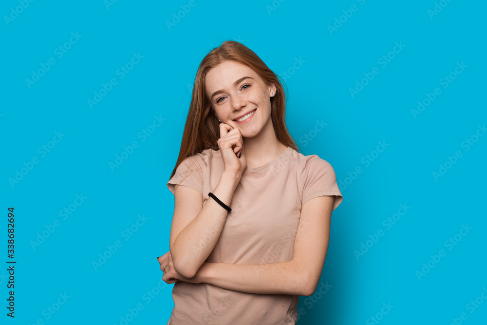 Ginger caucasian lady with freckles touching her chin and smile on a blue wall at studio Ginger caucasian lady with freckles touching her chin and smile on a blue wall at studio