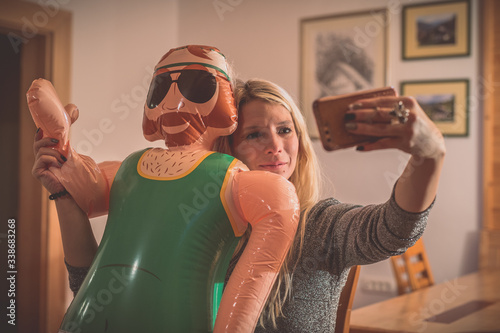 A cute blonde woman sending a message on a telephone while holding an inflatable male doll in a romantic way. Spending the holidays with fake friends. photo