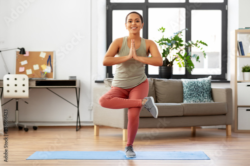 fitness, meditation and healthy lifestyle concept - happy smiling young african american woman doing yoga tree pose at home