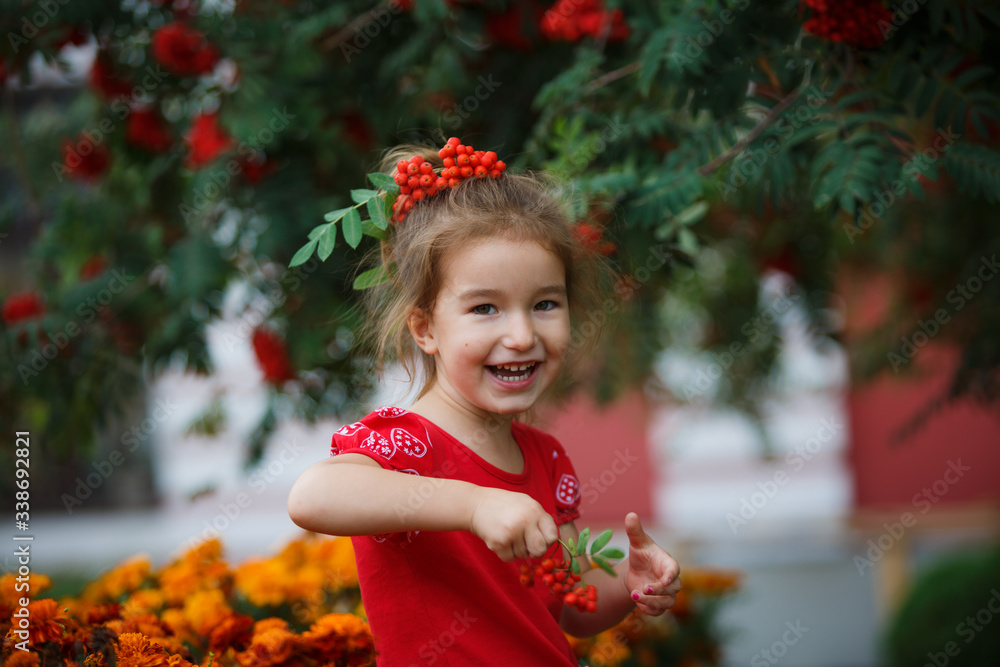 A little cheerful girl with Rowan berries on her head in a red dress. A girl in a Bush of ripe red Rowan laughs, mischievous and has fun.