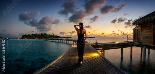 Eine attraktive Frau in schwarzem Kleid läuft über einen Holzsteg zu einer tropischen Insel auf den Malediven bei Sonnenuntergang