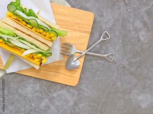 Fresh vegetable corn cheese with onion and cucumber sandwich. Baking paper on wood grain cutting board. Knife and fork. Close-up.Grey background 