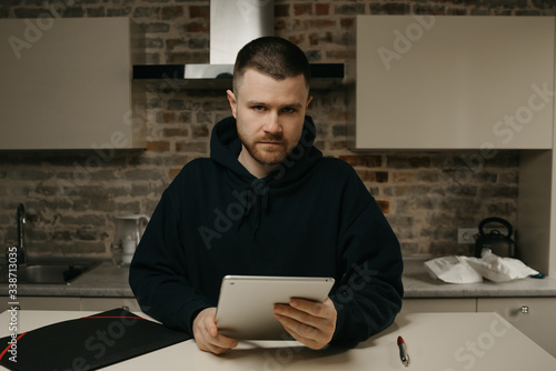 Remote work. A man works remotely holding his tablet. A businessman with a beard working right at his kitchen.