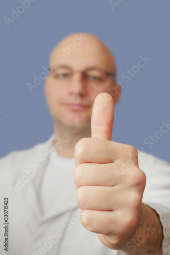 Thumbs up in focus, bald doctor with glasses in white coat out of focus, Blue background, Concept good and positeve news. photo