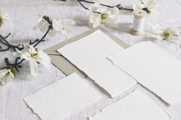 Wedding stationery set. Mock-up scene with blank paper greeting cards, envelope on linen tablecloth background with white magnolia stellata tree branches and ribbon. Feminine still life.