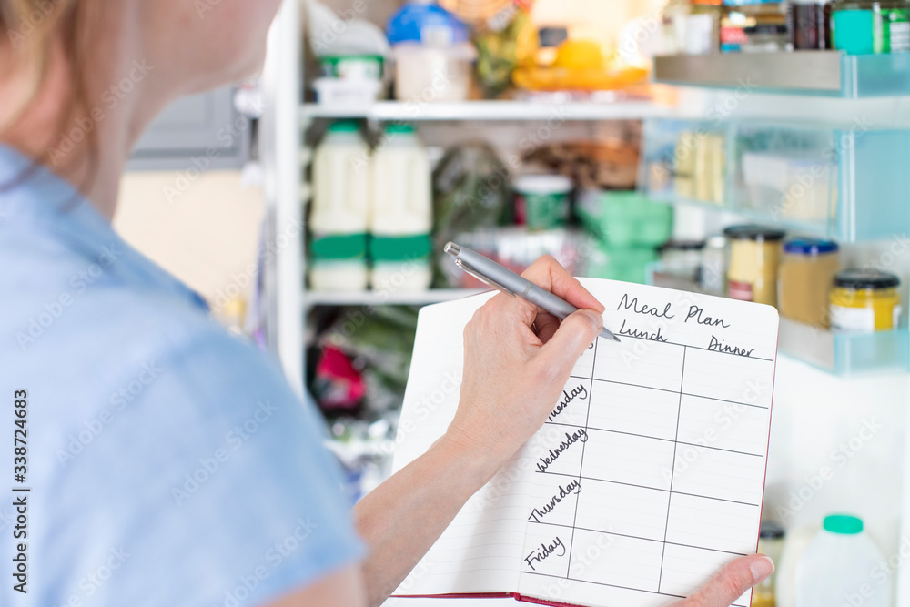 Woman Standing In Front Of Refrigerator In Kitchen With Notebook Writing Weekly Meal Plan