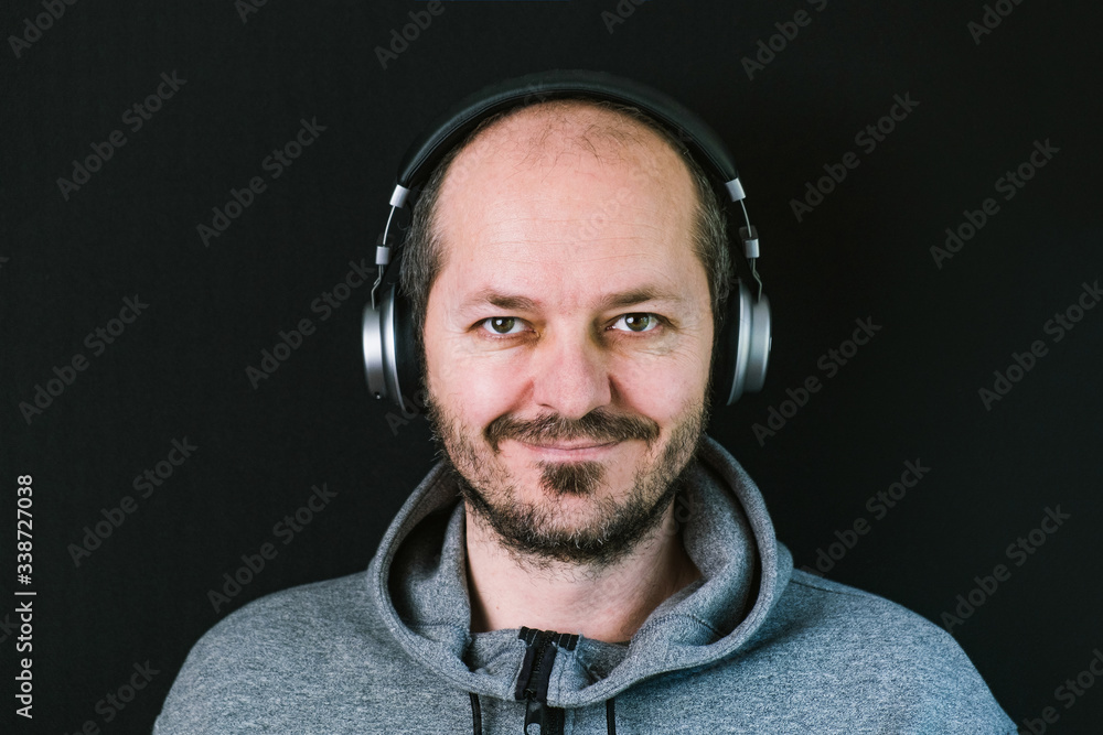 Bearded relaxed man in hoodie standing against black background with headphones, listening music, and smiling head and shoulders
Bearded relaxed man in hoodie standing against black background with headphones, listening music, and smiling head and shoulders