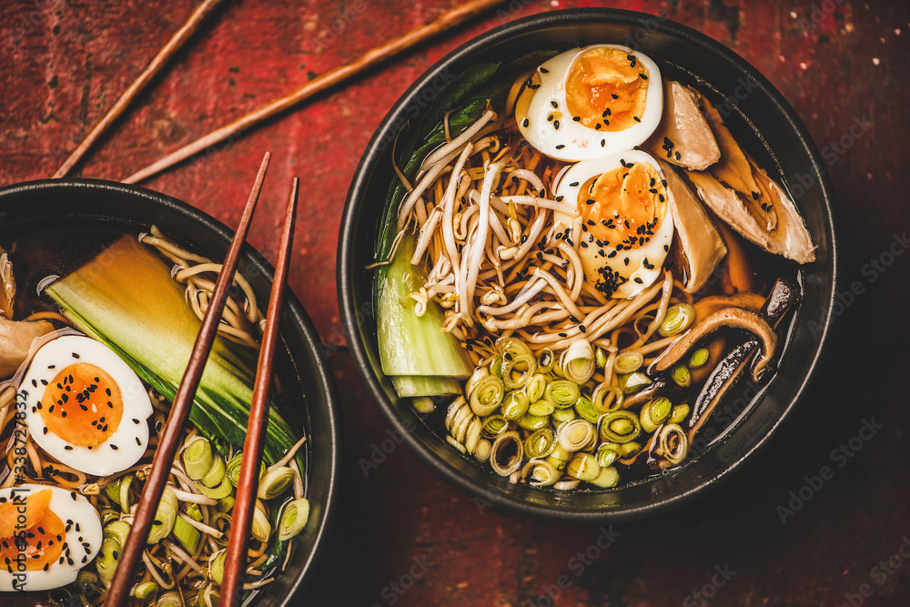 Flat-lay of traditional japanese Ramen soup with chicken meat, eggs, shiitake mushrooms in black bowls and bamboo chopsticks over dark red wooden table background, top view. Japanese cuisine concept Flat-lay of traditional japanese Ramen soup with chicken meat, eggs, shiitake mushrooms in black bowls and bamboo chopsticks over dark red wooden table background, top view. Japanese cuisine concept