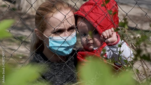 Woman in medical mask with small child isolated from society. Infected girl  isolated behind a grid.