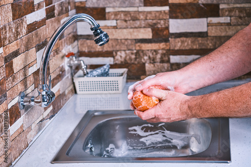 Man washing food due to coronavirus. Cleaning a tangerine to remove viruses. Metalic sink and soap to clean fruit.