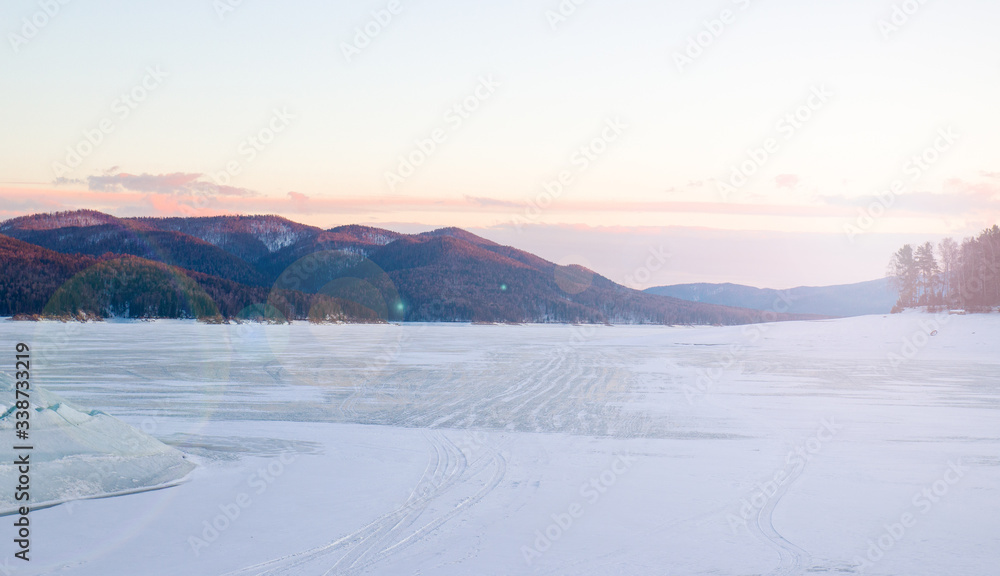 Awe winter landscape with a snowy frozen lake and mountains covered with coniferous forest at sunset with sun glare