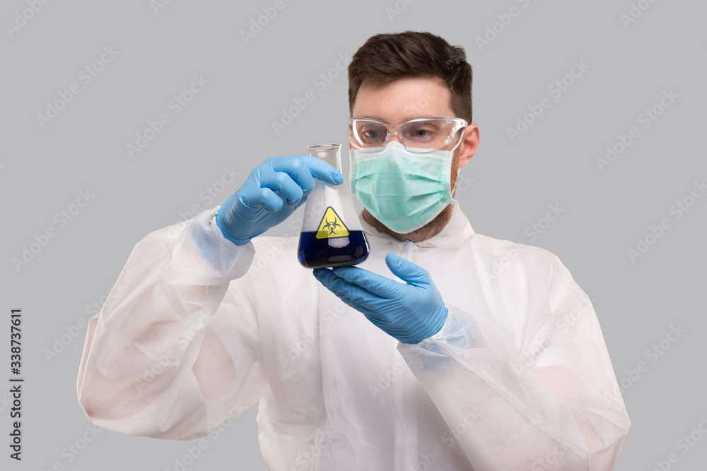 Male Laboratory Worker in Chemical Suit, Wearing Medical Mask and Glasses Watching Flask with Blue Liquid Biohazard Sign. Science, Medical, Virus Concept. Blue Liquid Male Laboratory Worker in Chemical Suit, Wearing Medical Mask and Glasses Watching Flask with Blue Liquid Biohazard Sign. Science, Medical, Virus Concept. Blue Liquid