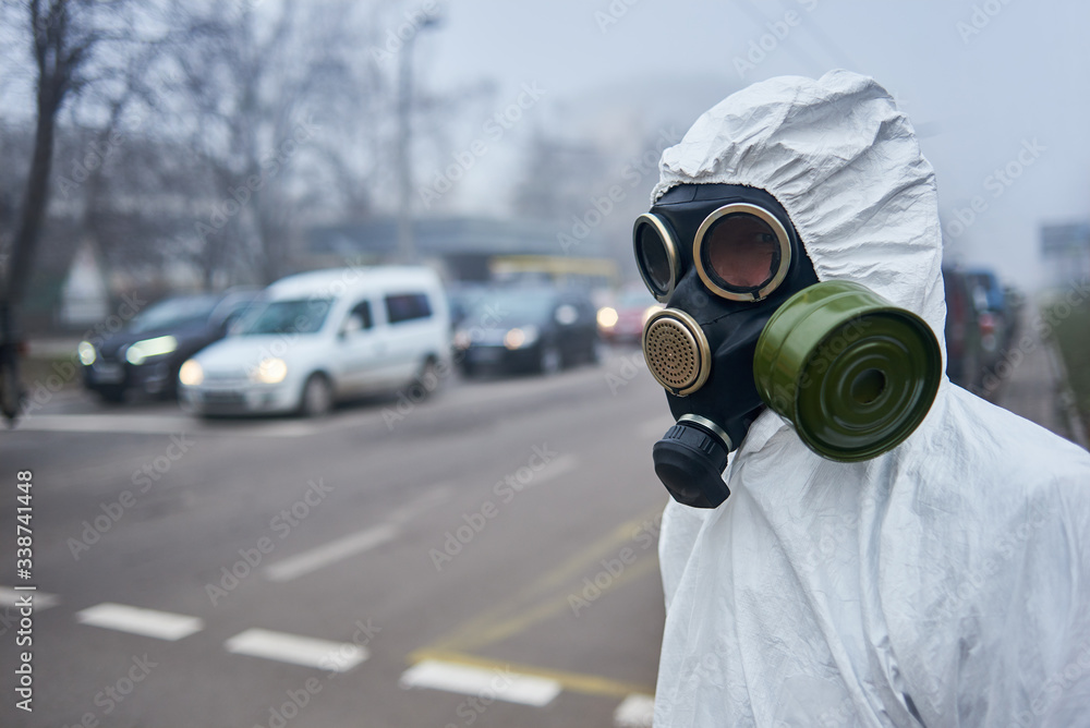 Selective focus of scientist wearing gas mask and protective costume looking at road with cars, waiting for green light. Close up of man posing near pedestrian crossing. Environmental disaster, fog. Selective focus of scientist wearing gas mask and protective costume looking at road with cars, waiting for green light. Close up of man posing near pedestrian crossing. Environmental disaster, fog.