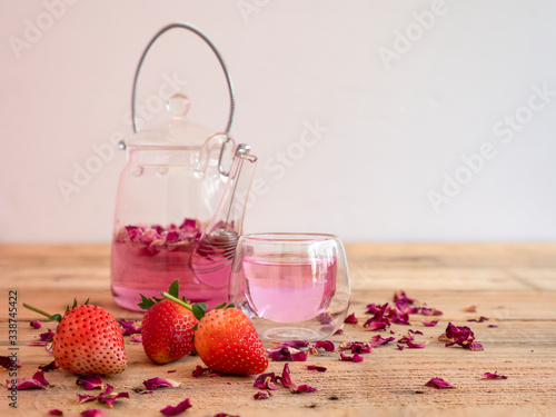 Pink rose petals infusion tea in a small clear drinking glass with glass kettle. Wooden rustic background.
