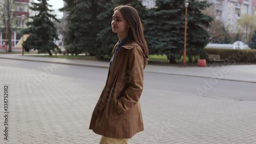 Young smiling woman walking in city. Side view of joyful female in casual clothes looking at camera and walking past columns of old building while strolling on street in sunny weather