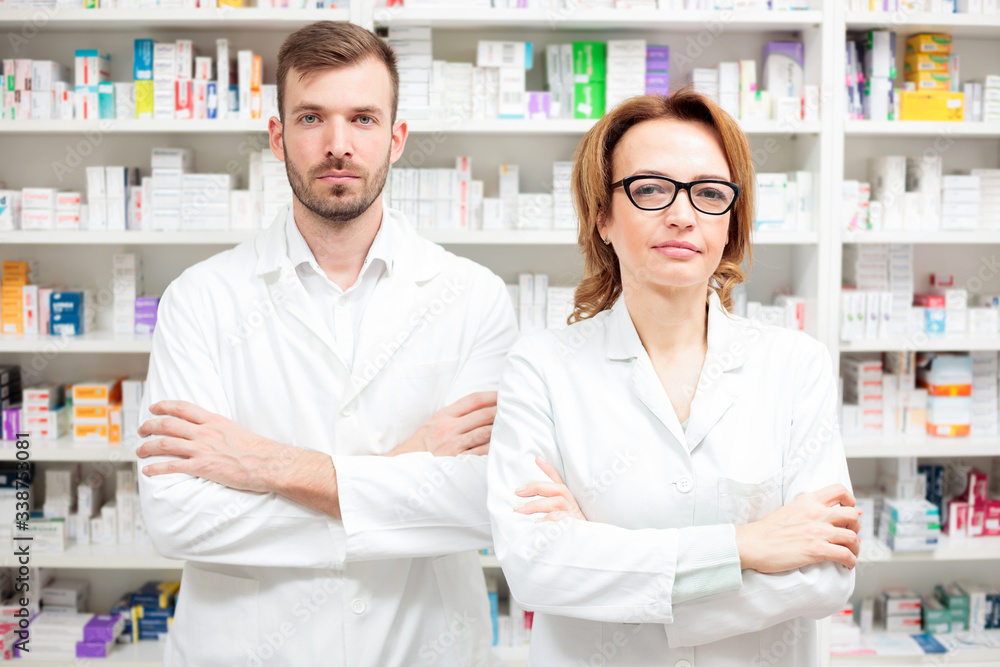 Two serious female and male pharmacists standing side by side in front of shelves, looking directly at camera with arms crossed on the chest. Healthcare and medicine concept