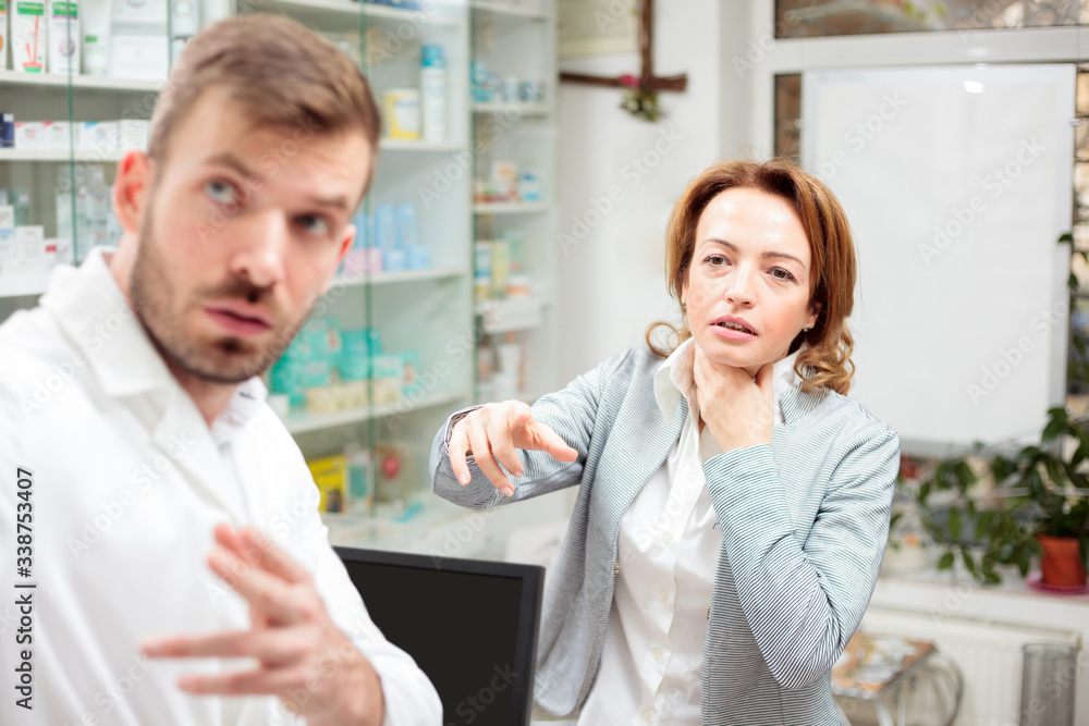 Serious mid adult woman with throat pain or thyroid gland problems seeking assistance in local pharmacy, young male pharmacist helping her. Healthcare and medicine concept Serious mid adult woman with throat pain or thyroid gland problems seeking assistance in local pharmacy, young male pharmacist helping her. Healthcare and medicine concept