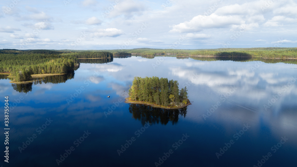 Beautiful island with green pine trees in the middle of  the large cold northern lake. Reflection of clouds on the surface of the lake.
