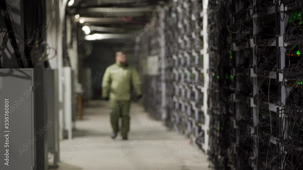 Data center diagnostics technician walks along the mining equipment racks in server room. Network administrator checks large computing cluster with network hardware. ASIC miners service engineer. Data center diagnostics technician walks along the mining equipment racks in server room. Network administrator checks large computing cluster with network hardware. ASIC miners service engineer.