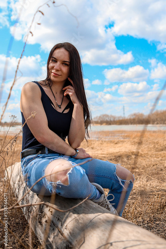 on the Bank of a small river, a log of a broken tree. A beautiful girl in blue jeans and a black t-shirt is sitting on it, enjoying the clean air.