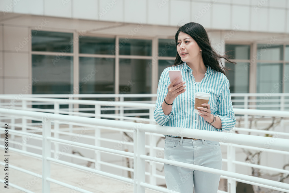young smiling asian chinese businesswoman walking on bridge outdoor in office work center using smartphone. happy relax lady enjoy break time texting message on mobile phone and holding cup of coffee