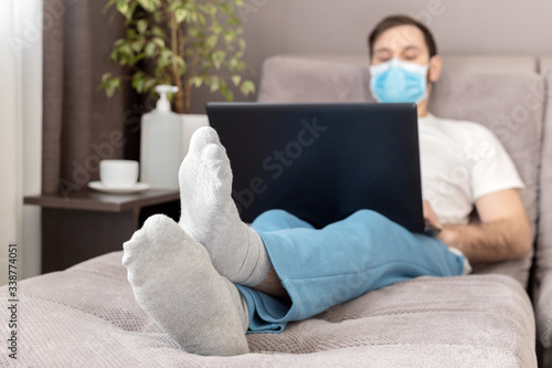 Young caucasian man working from home wearing protective mask using laptop and internet. Cozy Home office, workplace on sofa during coronavirus pandemic, covid 19 quarantine. Remote work, freelancer photo