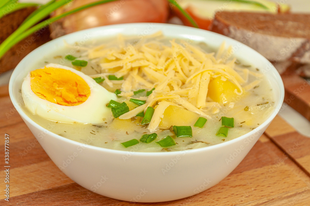 A white plate of cheese and potato onion soup with grated cheese poured on top and half a boiled egg on a cutting board against the background of rye bread and onions.