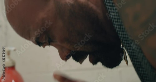 Bald, tan, muscular, bearded man splashes cold water on his face.