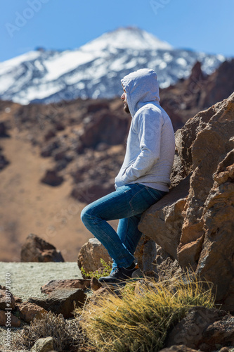 A young man with a hood sitting on the rocks while looking at the top of Teide mountain in Tenerife island, Spain