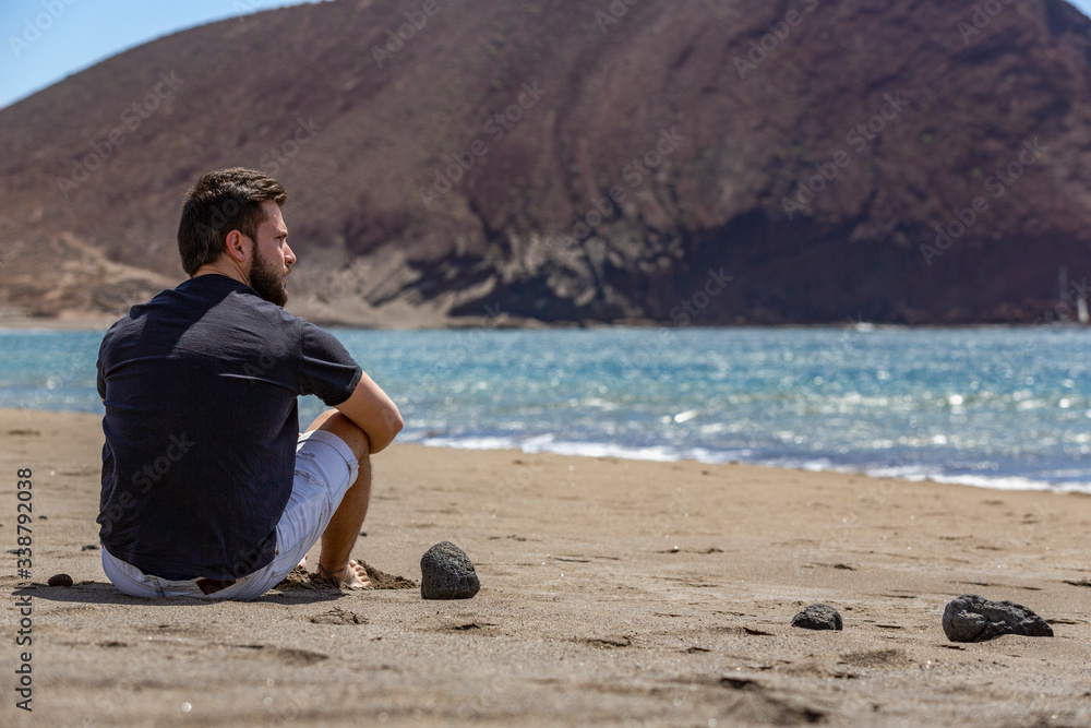 A young handsome man sitting on the sand in a beach while looking at the sea, in Tenerife, Canary Islands, Spain (closer view)