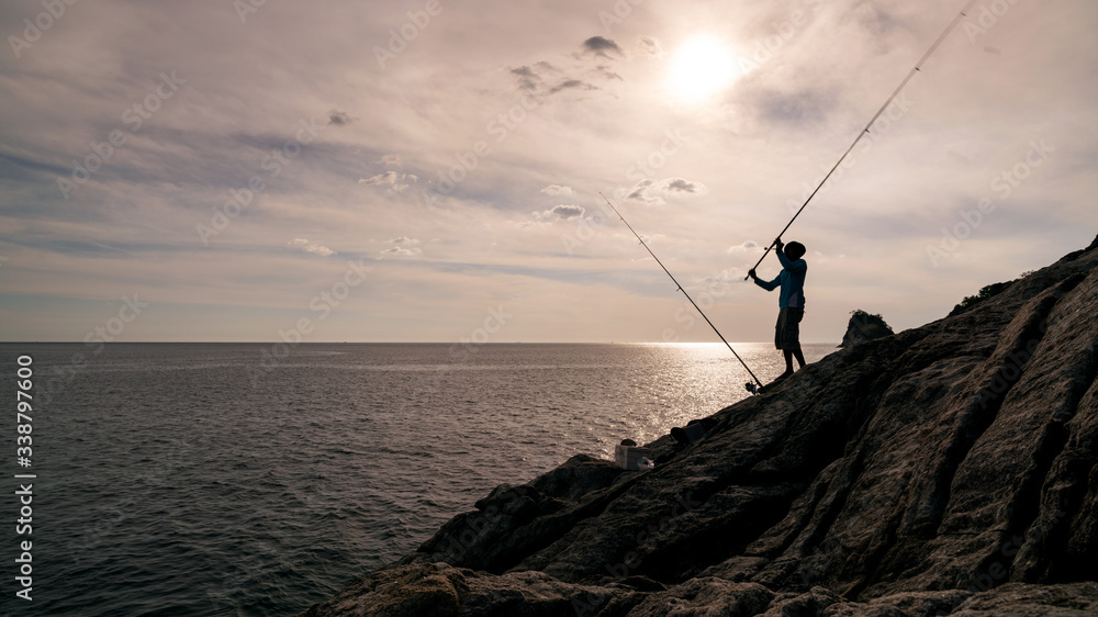 Silhouette of young man fishing on the rocks holding fishing rod in phuket Thailand.