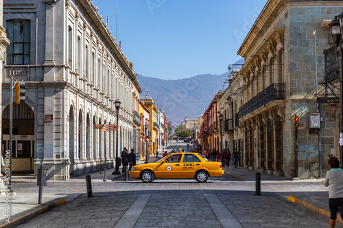 A yellow taxi cab drives throught the road near the main street in Oaxaca de Juárez, Mexico. Some people is walking by the streets in the morning