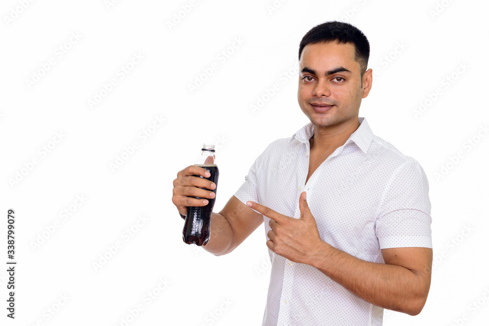 Studio shot of young handsome Indian man holding bottle of soda and pointing finger Studio shot of young handsome Indian man holding bottle of soda and pointing finger