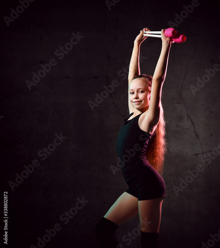 Young smiling girl gymnast in black sport body and uppers standing and holding two pink gymnastic maces in raised hands