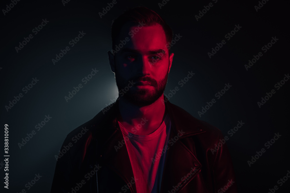 Portrait of young caucasian man with beard in leather jacket illuminated neon light looking at camera. Dark smoke studio background. Portrait of young caucasian man with beard in leather jacket illuminated neon light looking at camera. Dark smoke studio background.