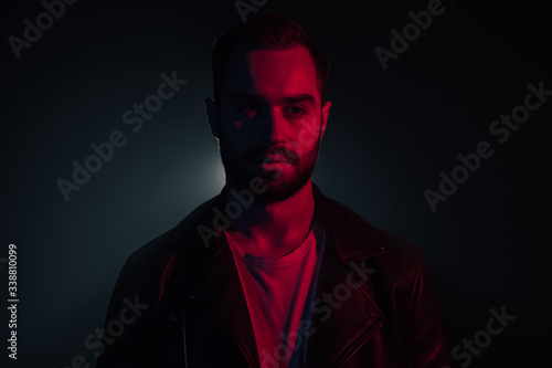 Portrait of young caucasian man with beard in leather jacket illuminated neon light looking at camera. Dark smoke studio background.