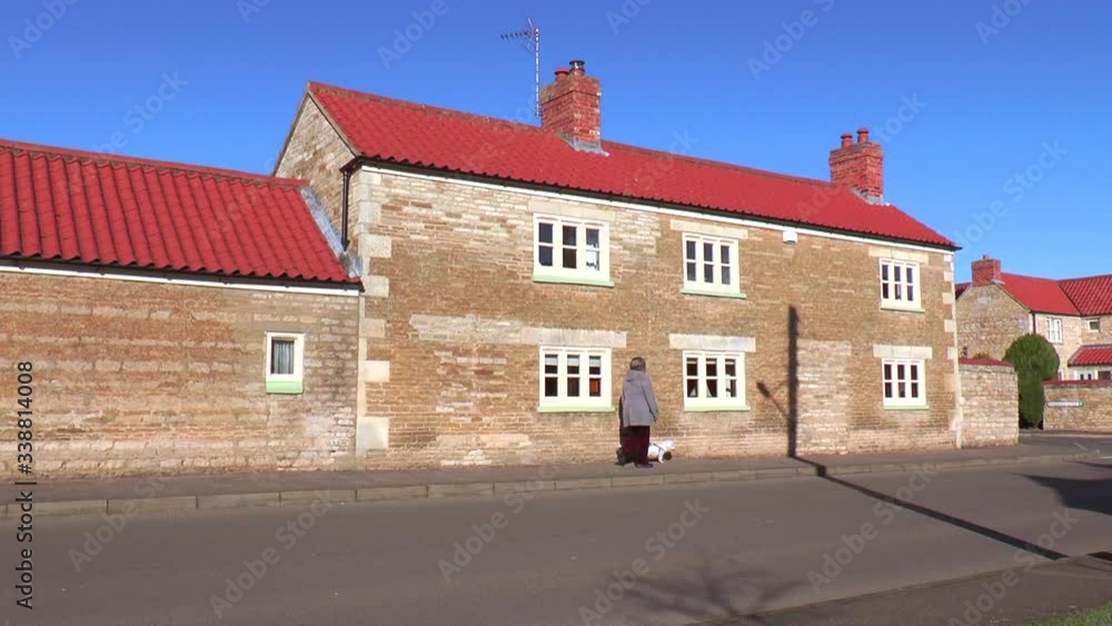 A mature woman walking her Bichon Frise pet dog on a footpath in an English village, in the warm autumn sunshine, past an old stone house against a clear blue sky. A mature woman walking her Bichon Frise pet dog on a footpath in an English village, in the warm autumn sunshine, past an old stone house against a clear blue sky.