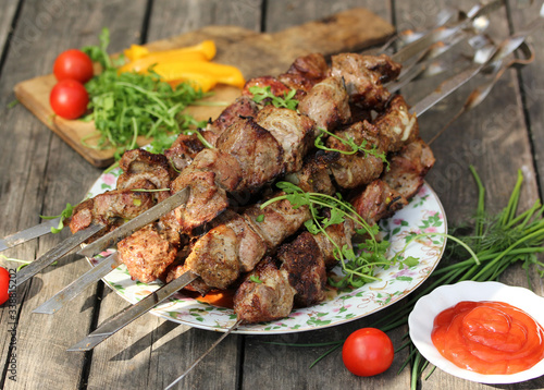 Grilled pork shashlik on skewers on a light plate on an old wooden background with ketchup in a bowl, tomatoes, peppers and herbs. Background image, copy space