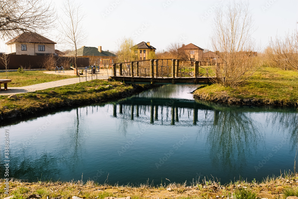 Blue pond with sky reflection and village houses.Wooden bridge over a small river or pond, side view. Pedestrian bridge over the river on