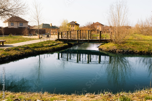 Obraz na płótnie Blue pond with sky reflection and village houses.Wooden bridge over a small river or pond, side view. Pedestrian bridge over the river on w salonie