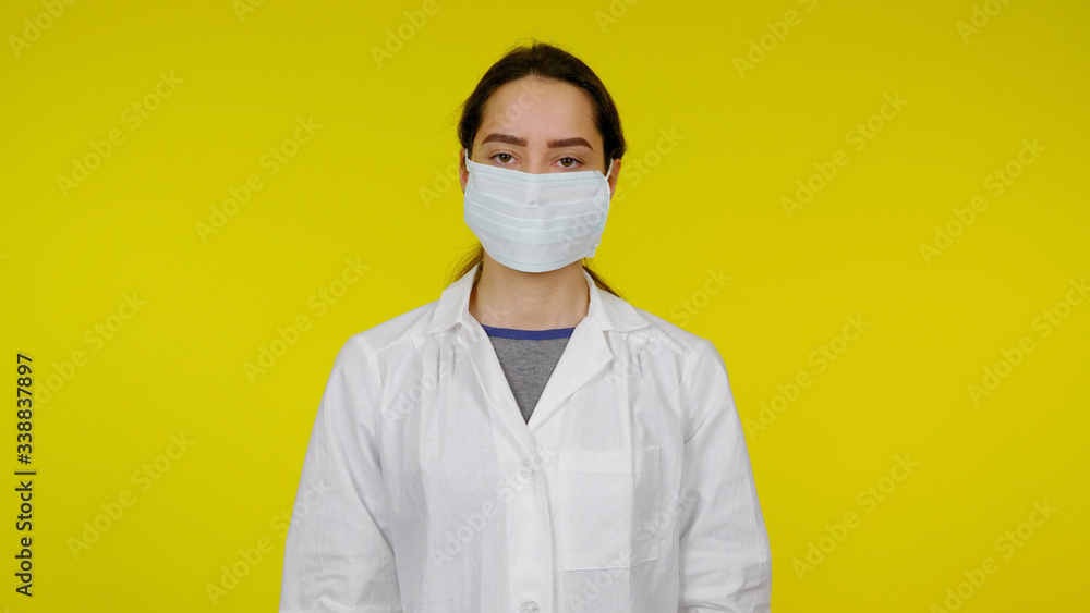 Young infectious disease doctor in a protective medical mask on her face looks at the camera. Girl in a white coat on a yellow background. Coronavirus, flu