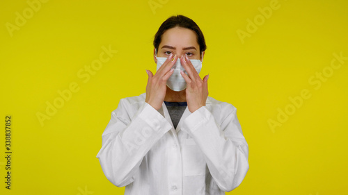 Young doctor infectious disease specialist adjusts a protective medical mask on her nose. Girl in a white coat against the yellow background puts on a mask on face, covering her nose and mouth
