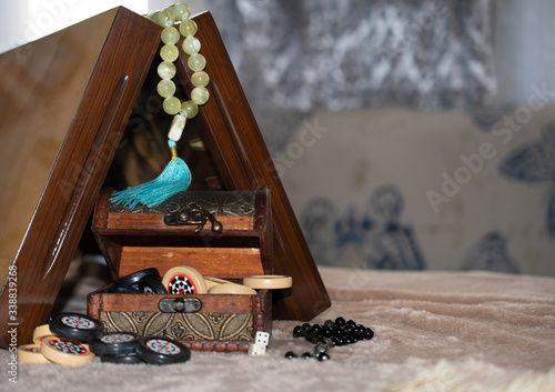 An old little chest is open and you can see black and white patterned backgammon chips, black opal rosaries on top, and next to them dice with numbers under the open playing Board and onyx rosaries 