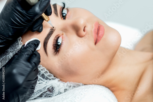 Professional cosmetologist hands are doing permanent makeup on eyebrows of young woman, close up.