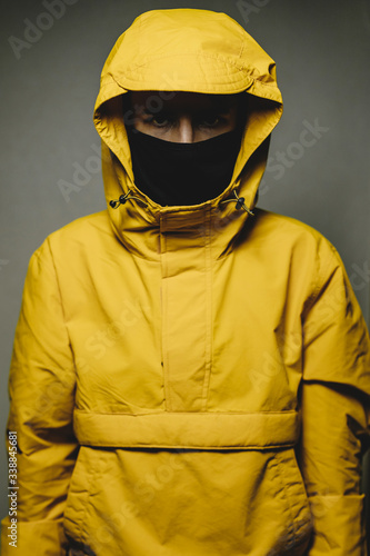 young skinny man wearing a yellow hooded jacket and black face mask with an aggressive serious look © Максим Галінский